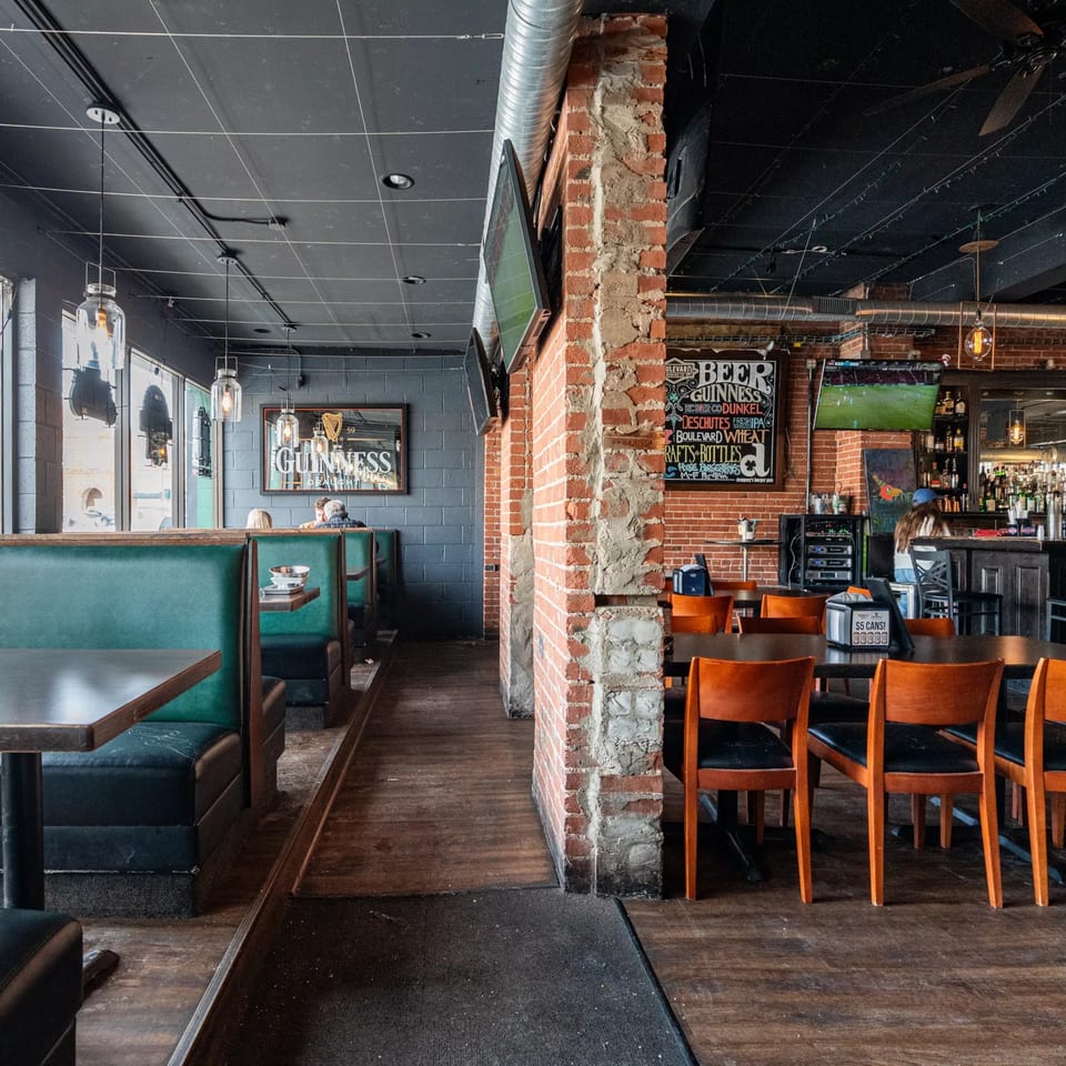 Interior of Dempsey Burger Bar, showing green booths, wooden tables, brick walls, and a bar area.