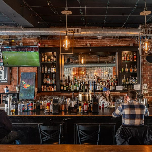 A bartender serves a customer at the Dempsey Burger Bar, with a TV showing soccer and a full liquor shelf.