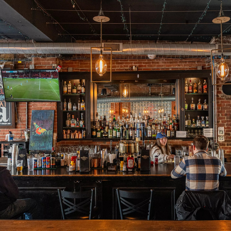 A bartender serves a customer at the Dempsey Burger Bar, with a TV showing soccer and a full liquor shelf.