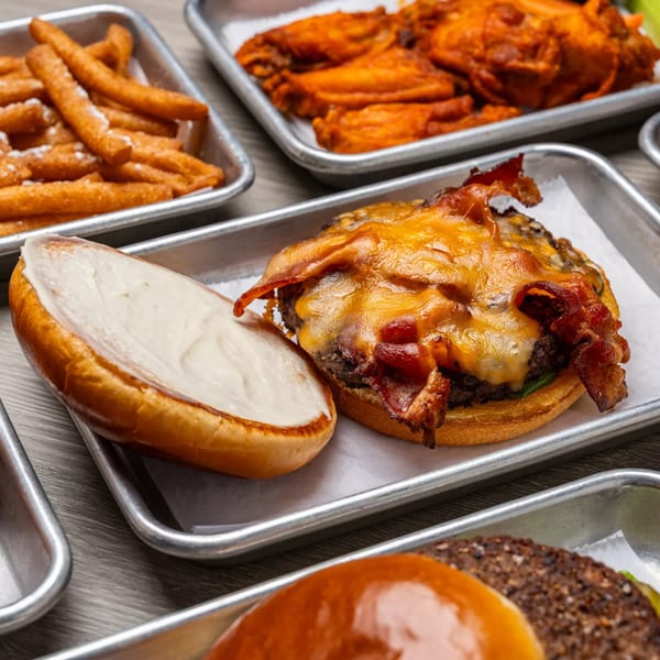 A Dempsey Burger Bar bacon cheeseburger with fries and chicken wings on metal trays.